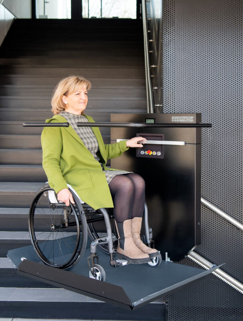 Frau sitzt in ihrem Rollstuhl und fährt mit dem Plattformlift die Treppe hinunter