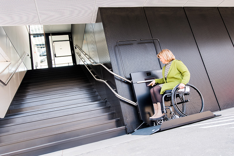 Frau sitzt mit Rollstuhl auf Plattformlift und startet fahrt die Treppe hinauf